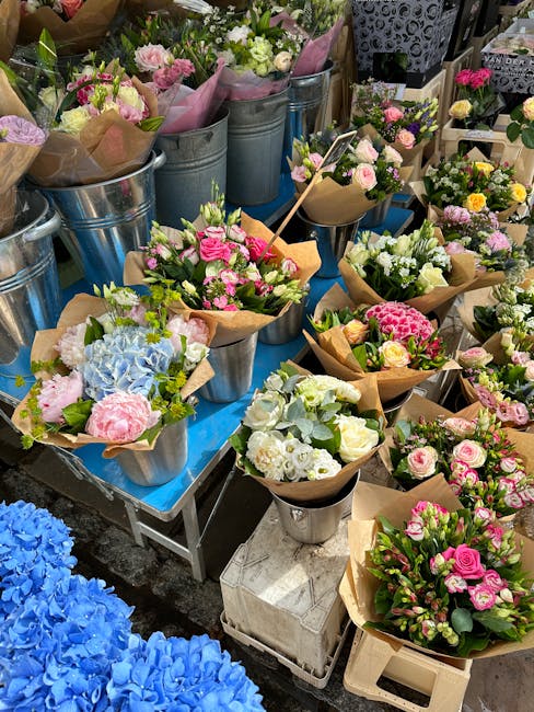 A display of freshly arranged floral bouquets and potted flower arrangements featuring pastel pink roses, white roses, blue hydrangeas, yellow and white spray roses, and mixed greenery, all wrapped in