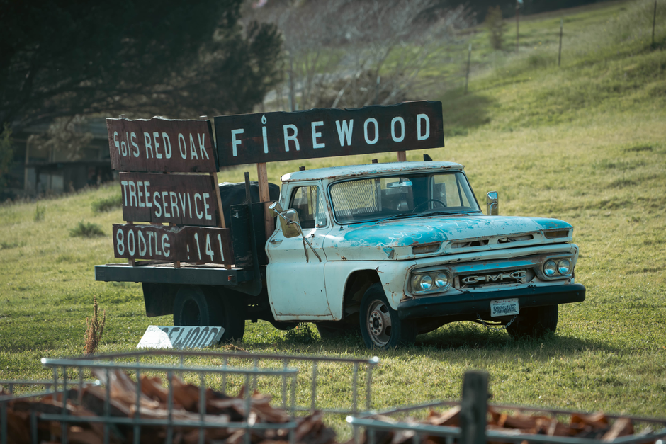 An outdoor scene featuring an old, weathered GMC pickup truck painted pale blue with rust patches, positioned on a grassy field. The truck's bed is loaded with large, flat signs advertising local serv