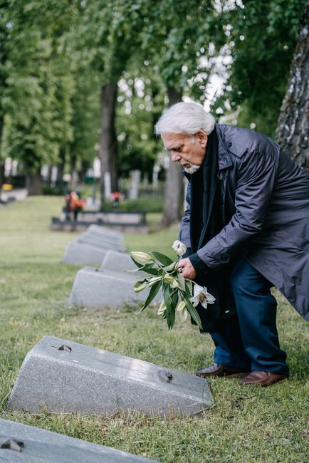 An elderly man with grey hair, dressed in a navy jacket and dark trousers, is placing a small floral bouquet on a gravestone in a lush, green cemetery. The bouquet consists of white lilies, roses, and
