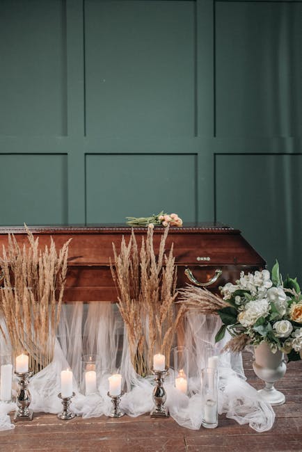 A wooden funeral casket resting on a table draped with white sheer fabric, surrounded by tall, beige pampas grass and several white and pale pink candles in silver holders. To the right, there is a la