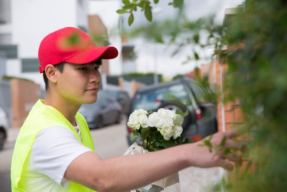 A young male flower delivery courier wearing a red cap and a neon yellow vest is outdoors, placing a fresh floral arrangement on a doorstep. The bouquet consists of white hydrangeas held in clear plas