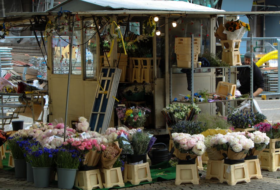 A flower stall set up outdoors under a canopy displays a variety of fresh flowers arranged in black buckets and wrapped bouquets, featuring pink, white, purple, and blue blooms. The flowers include ro