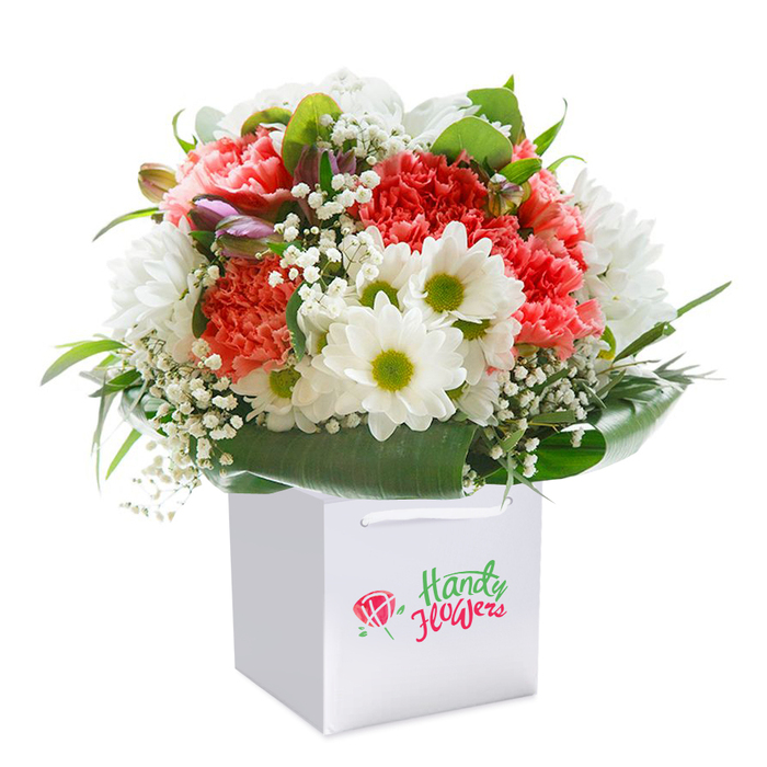 A stunning composition worthy of Old Oak Common's vibrant community, this floral arrangement flourishes inside a contemporary white gift bag, elegantly set against a luminous white background. A symphony of coral-pink, peach, and ruby red carnations forms the heart of the bouquet, their abundant, frilled petals offering softness and texture as layered and inviting as the green expanses near Willesden Junction. Interspersed, snowy white daisies capture attention, each bright yellow-green center sparking with vitality, reminiscent of children's laughter across local playgrounds. Wisps of ethereal, white baby's breath float easily through the blooms, their light touch introducing a sense of poetry and making every stem glow with understated elegance. A cluster of unopened purple buds nestles discreetly on one side-subtle, promising, intriguing. Surrounding the flowers, a sweep of deep emerald leaves and glossy foliage creates a lush, sculpted collar, neatly containing the exuberance above and lending structure and lasting freshness. The white bag's sturdy ribbon handles are both practical and stylish, designed for convenient delivery-ideal for a birthday, thank you, or simply bringing a touch of Old Oak Common's charm indoors. Balanced, professional lighting amplifies the arrangement's vividness and crispness, capturing the meticulous craft and local expertise that define the area's best floristry. This bouquet shines as a testament to comfort, celebration, and the power of a thoughtfully delivered floral gesture.