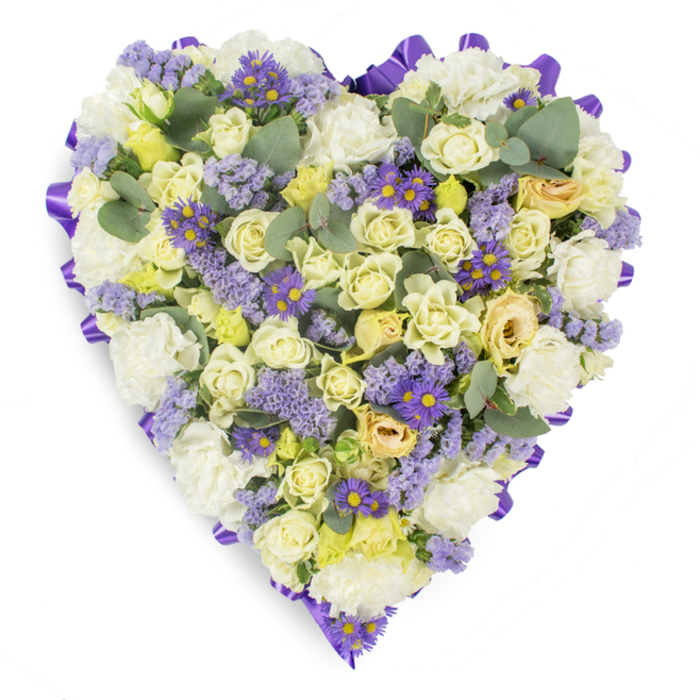 Presented against a spotless white backdrop, this heart-shaped memorial arrangement reads as a measured, comforting composition of cream, white and pale yellow florals, edged with a band of deep purple satin ribbon that defines its silhouette with quiet formality. The principal elements are tightly arranged cream and pale yellow roses, their plush, concentric petals creating gentle tonal shifts from ivory to buttery hues and occasional hints of light green at the bud bases. Nestled among these are plentiful white carnations whose ruffled faces add body and a timeless sympathy language to the piece. Accents of lavender statice introduce an airy, feathery texture while bright purple, aster-like flowers with vivid yellow centres provide small, uplifting points of contrast across the heart. Scattered rounded eucalyptus leaves in a blue-green hue punctuate the display, offering sculptural foliage and a restrained, silvery coolness that visually calms the dense floral mass. The satin ribbon around the edge is looped and folded to create a continuous, polished border, lending the tribute a finished, ceremonial feel appropriate for funeral chapels, wakes, or graveside services near Old Oak Common, North Acton and the developing Wilton Crescent area. The soft, neutral lighting accentuates petal surfaces and the arrangement's layered depth, while the overall mood remains one of solace, respect and lasting remembrance - a floral design intended to comfort mourners and honour the memory of a loved one.